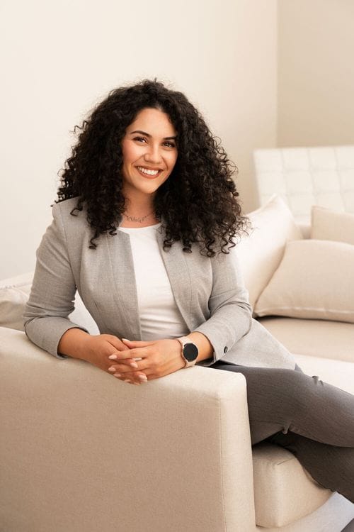 A woman with curly hair smiling and sitting on a cream sofa in a modern living room.